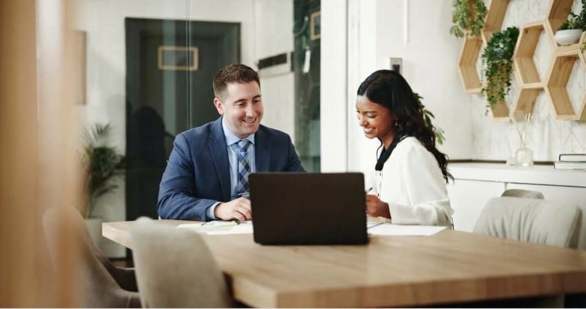 Two professionals in business attire having a meeting at a wooden desk with a laptop in a modern office space